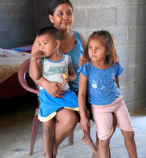 A smiling woman holds her son and daughter close outside a concrete-block house.