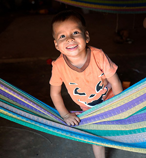 A boy inside a dirt-floor home stands behind a colorful hammock and smiles.
