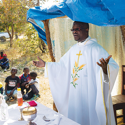 A priest in white vestments lifts his hands while celebrating Mass outdoors.