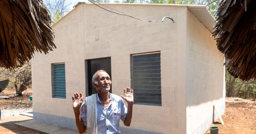 A man smiles and raises his hands toward heaven as he stands in front of a concrete-block home.