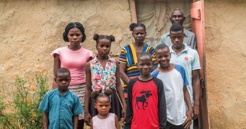 A mother, father, and seven of their eight children stand in front of their crumbling home.