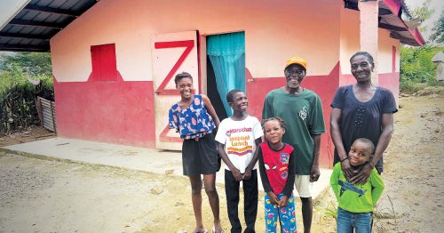 A family smiling and laughing outside a brightly colored concrete-block home.