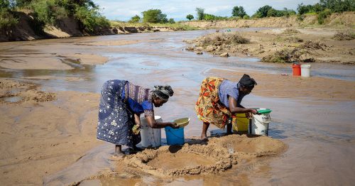 Two Malawian women bend over to collect water from a river.