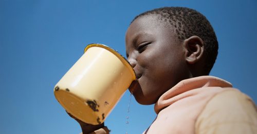 A girl drinks water from a yellow tin cup.