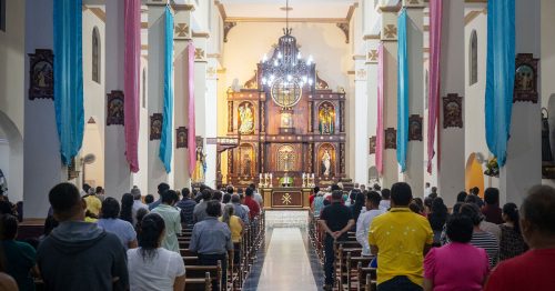 Seen from the rear, the inside of a Catholic church filled with people standing in front of their pews.