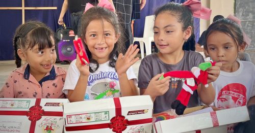 A young girl with a box in her lap joyfully plays a small tambourine as her friends look on.