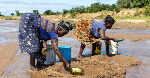 Two women scoop water from a shallow and muddy stream using bowls and buckets.