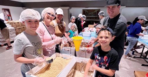 Parishioners of all ages, wearing hair covers, stand over boxes of rice at a food-packing event.