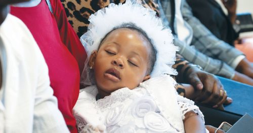 A female toddler in a white outfit sleeps peacefully in a woman’s arms.