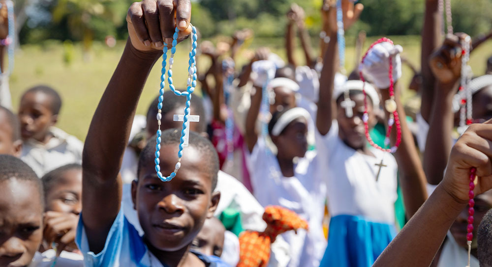 Many children, who are standing outside, hold up rosaries high above their heads.