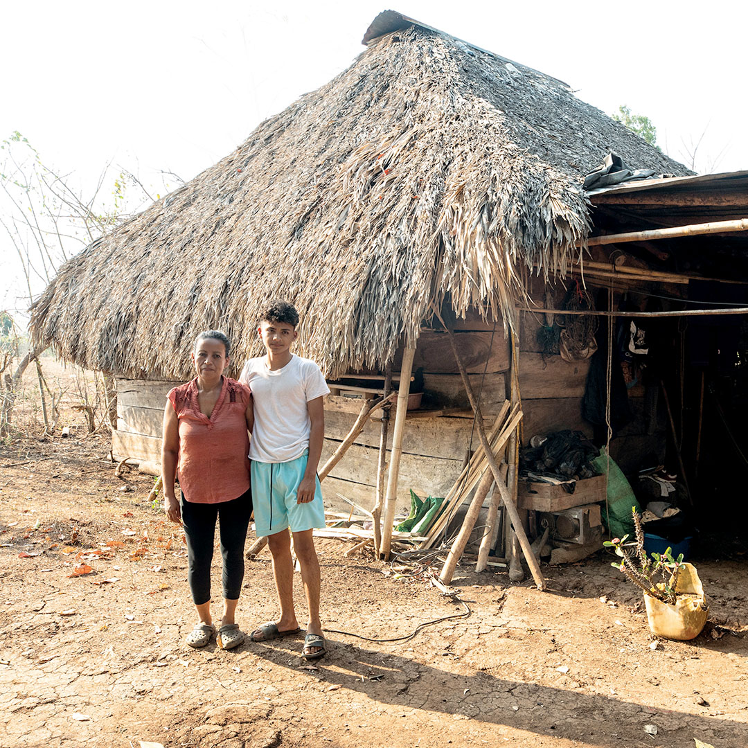 A mother and son stand in front of a home made of scrap wood, metal and palm fronds.
