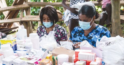 Two nurses in masks sit behind a table filled with medications.