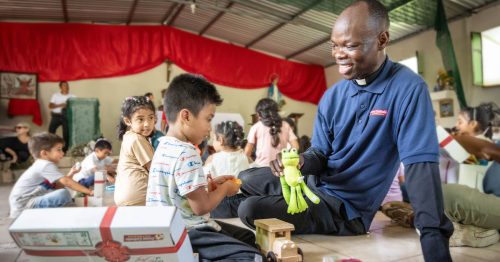 A young boy and a Catholic priest sit on the ground while the boy opens toys from a gift box.