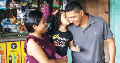 A woman holds her daughter, who leans over to give her father a kiss.
