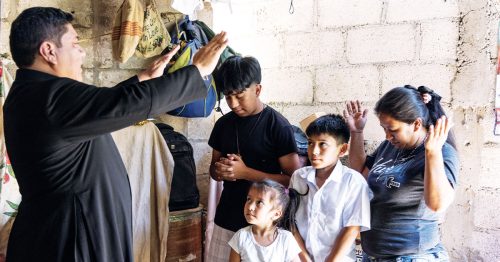 A family of four prays in their home while a priest raises his hands in blessing over them.