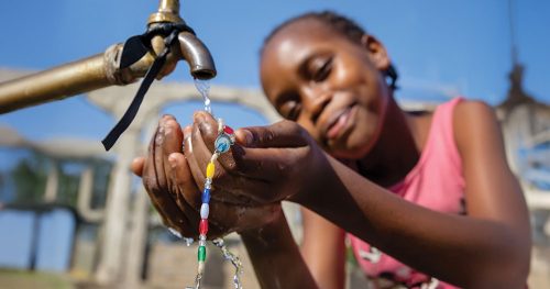 A little girl with a rosary puts her hands under an outdoor faucet with flowing water.