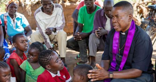 A Catholic priest sits and talks to a group of children and adults.