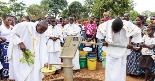 A priest in white vestments blesses a hand pump well while an altar boy pumps water.