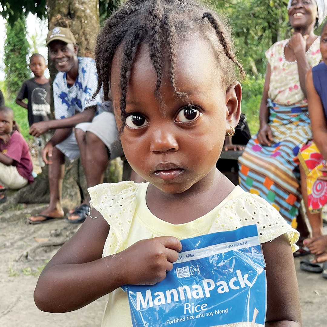 A young girl holds a Vitafood meal pack while caregivers and other children in the background look on.