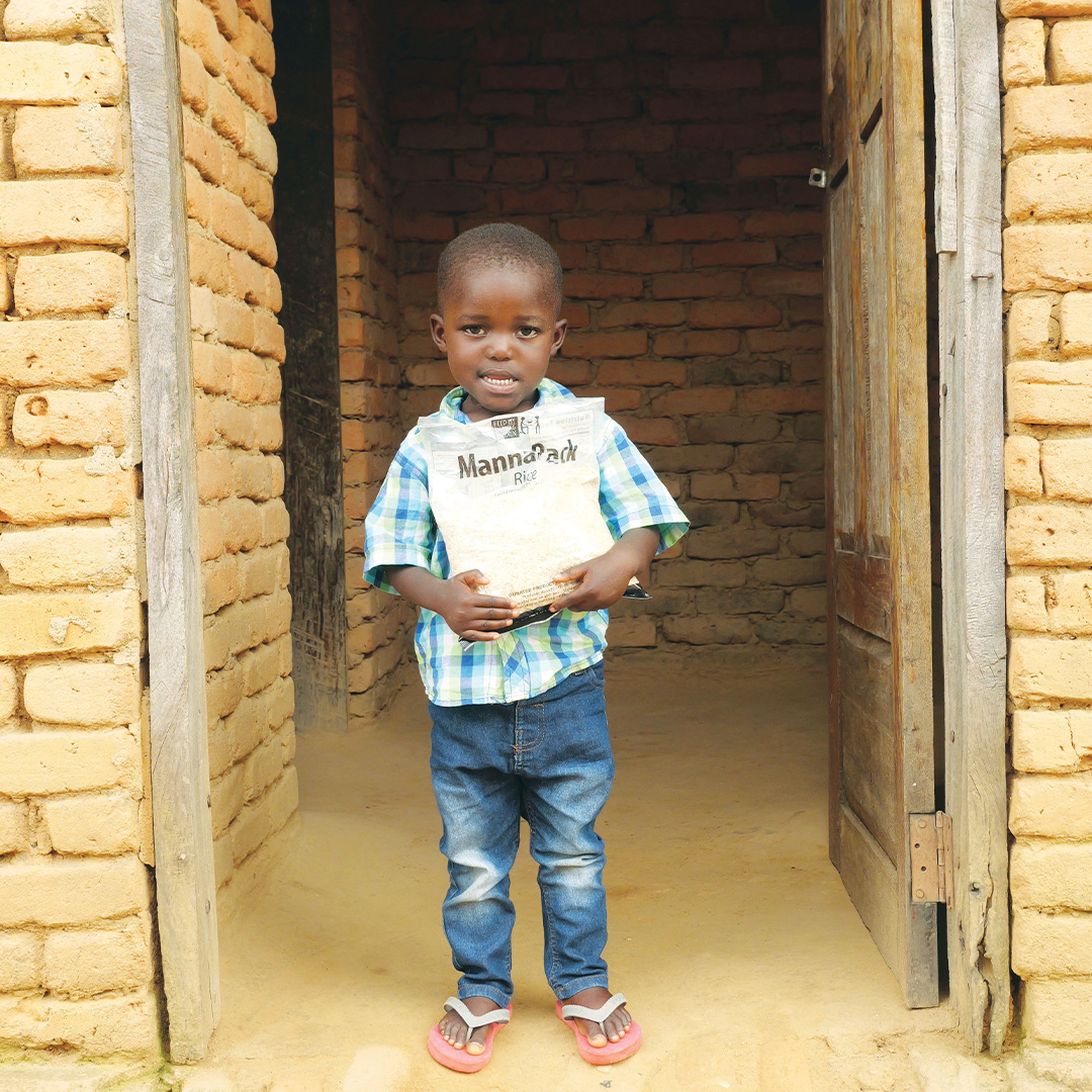 A small boy stands in the doorway of a home in Malawi, holding a Vitafood meal pack.
