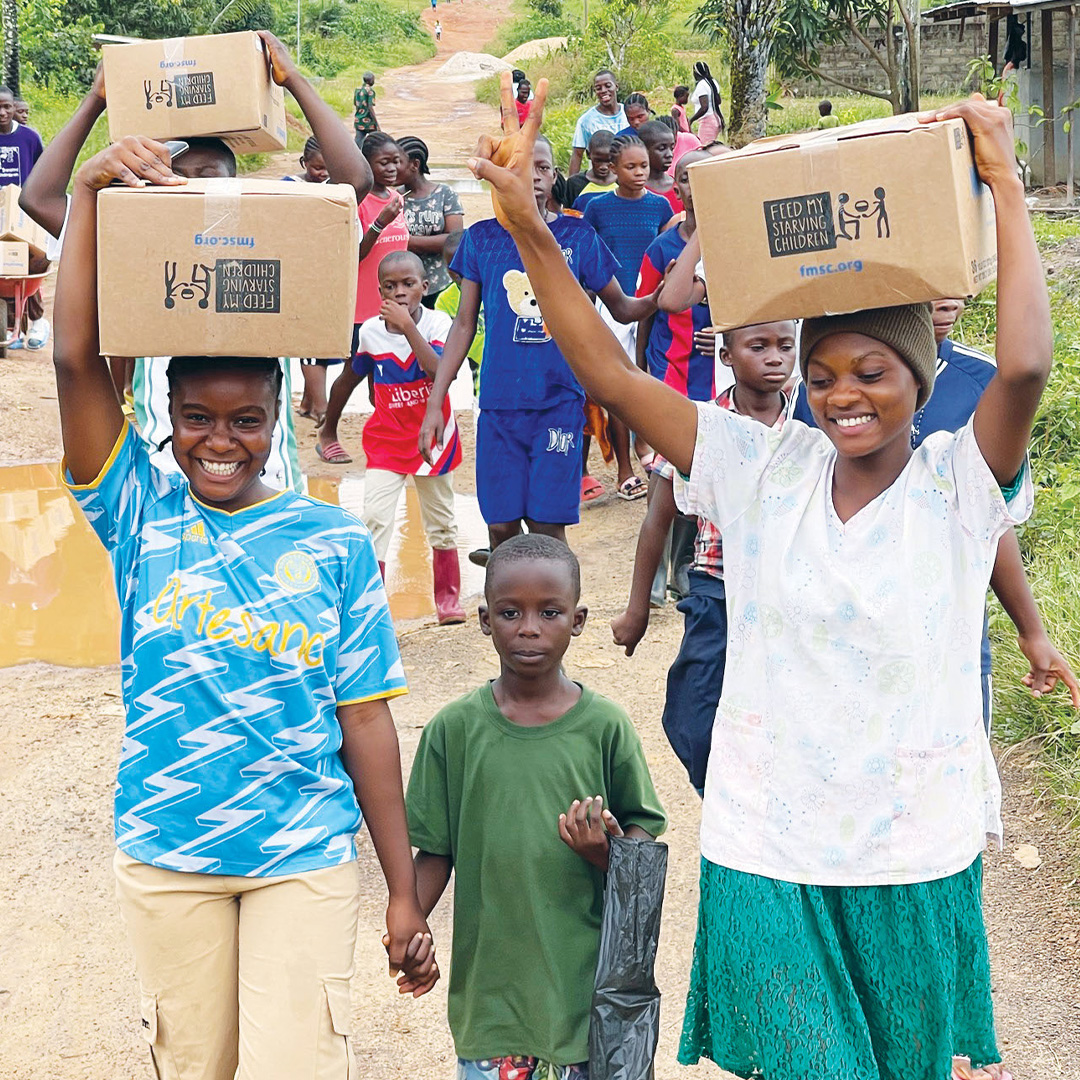 A group of children and young people carry cardboard boxes of Vitafood down a dirt road in Liberia, smiling and laughing.