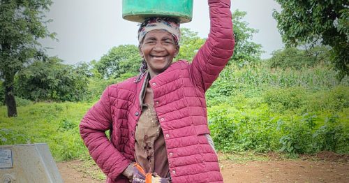 A woman stands at a water pump, holding a bucket on her head.