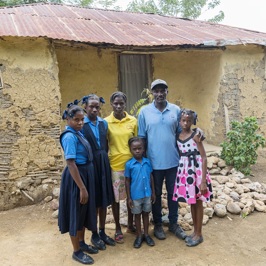 A family of six stands in front of a crumbling house.