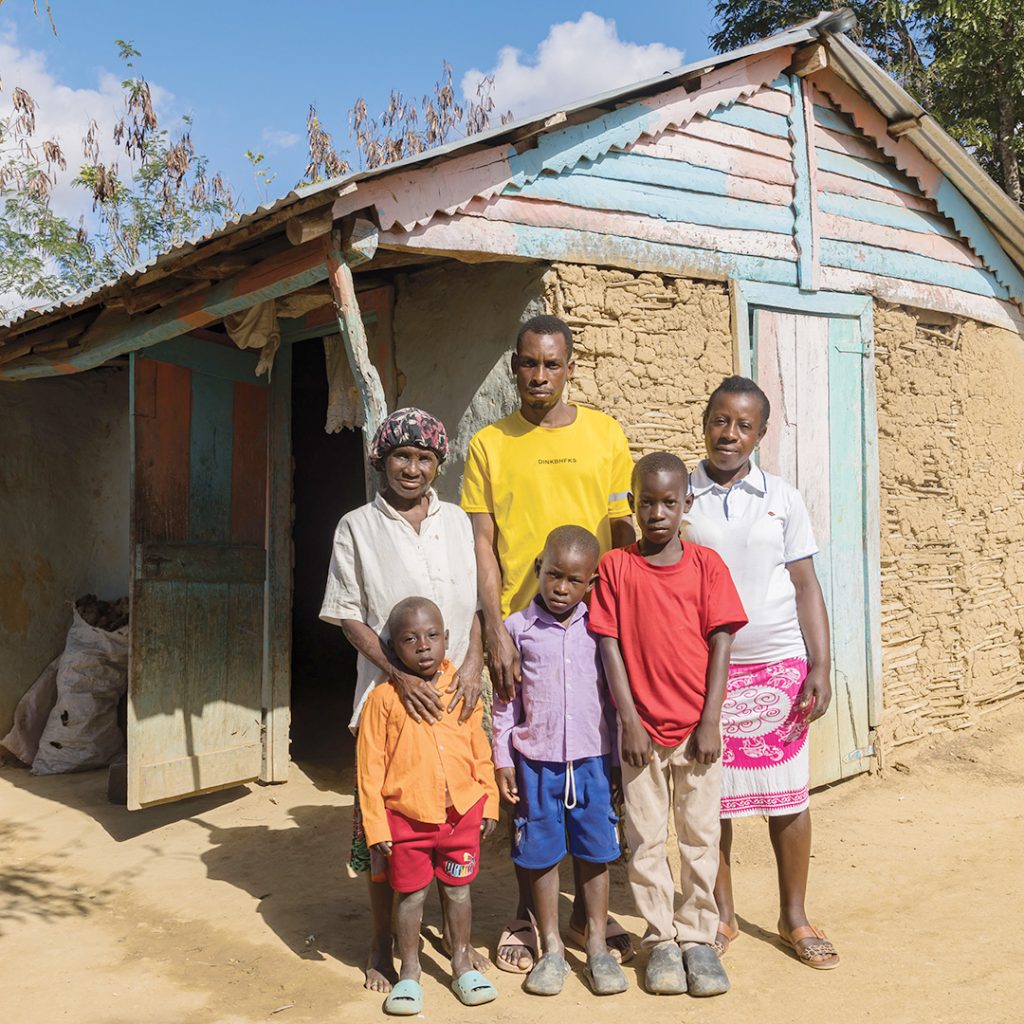 A family with young children stands in front of a small mud house.