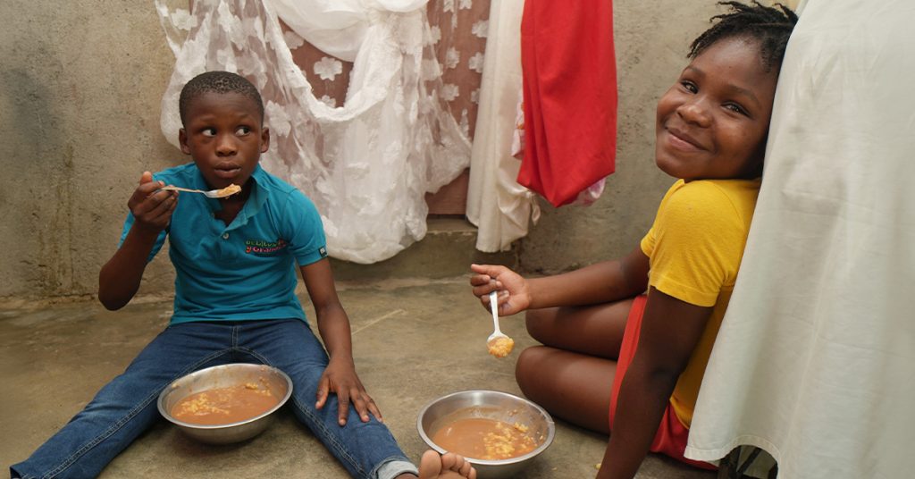 Two children seated on the dirt floor of their home eat soup from metal bowls.