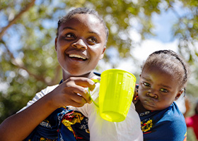 A young girl carries her sister on her back while holding a cup of water.
