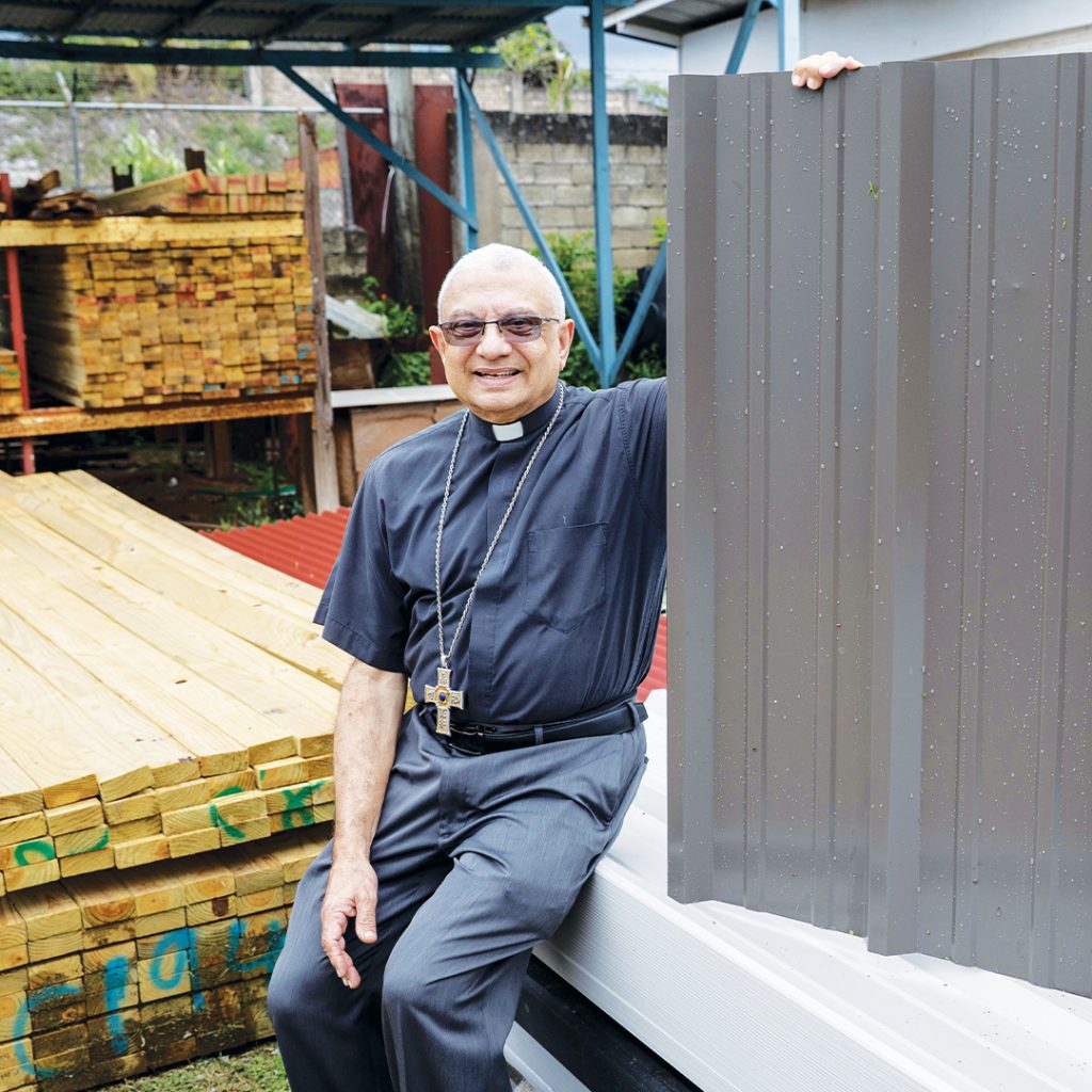 A Catholic bishop holds up a galvanized roof panel while sitting near stacks of lumber and other construction materials.