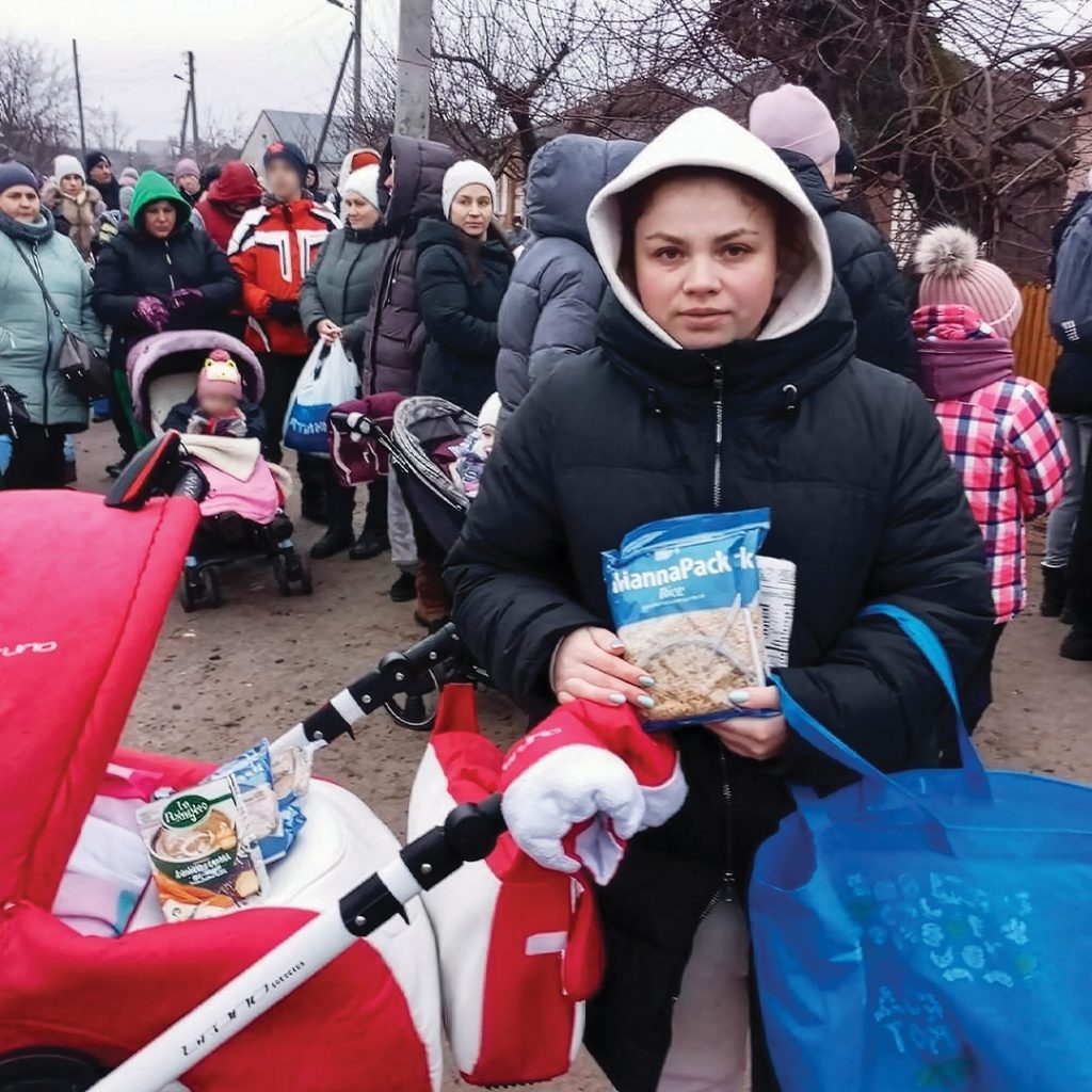 A woman with a baby stroller holds a pack of rice and beans as a crowd of people waits in the background.