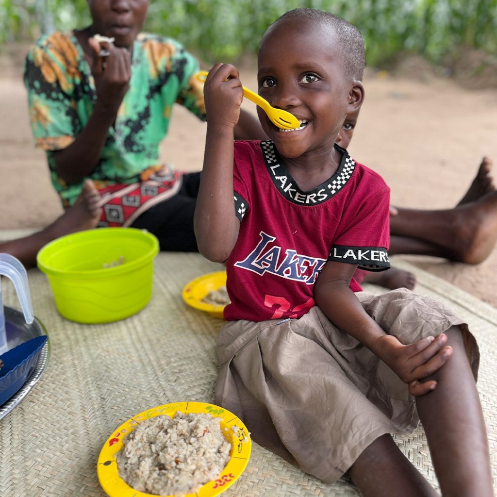 Three children sit on a woven mat outdoors eating rice from colorful bowls.