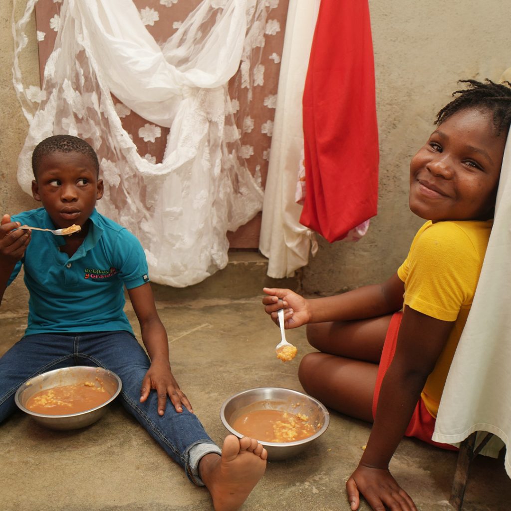 Two children sit on the dirt floor of their small home eating bowls of food.