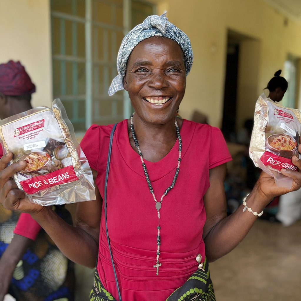 A smiling woman wearing traditional clothing and a rosary necklace holds up two Vitafood meal packs.
