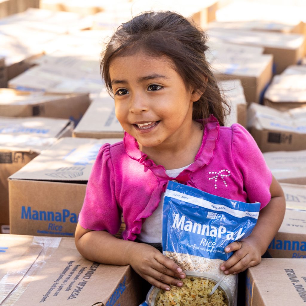 A little girl in a pink top stands among boxes of Vitafood packs, holding one pack in her hands.