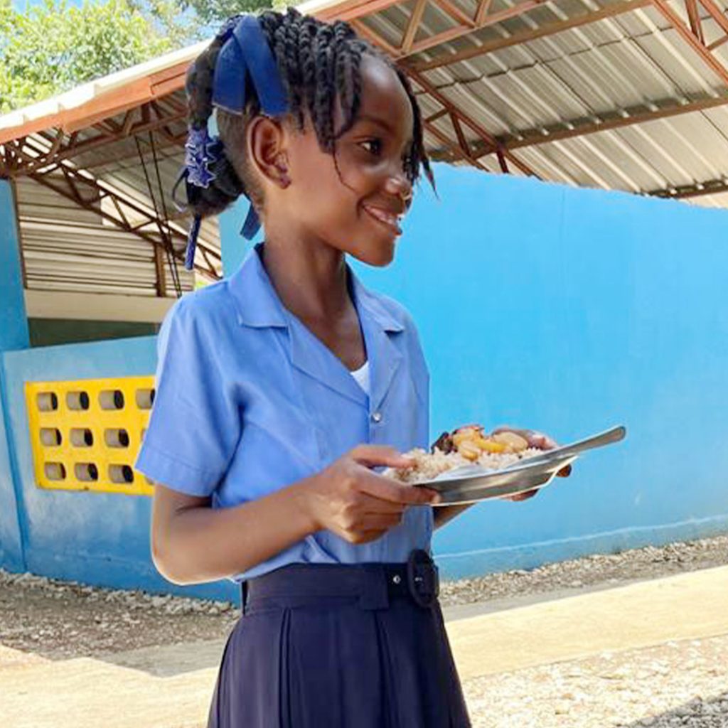A young girl in a school uniform holds a plate of food outside her open-air classroom in Haiti.
