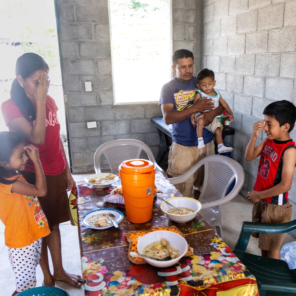 A family stands and prays around a small dining table set with bowls of food.