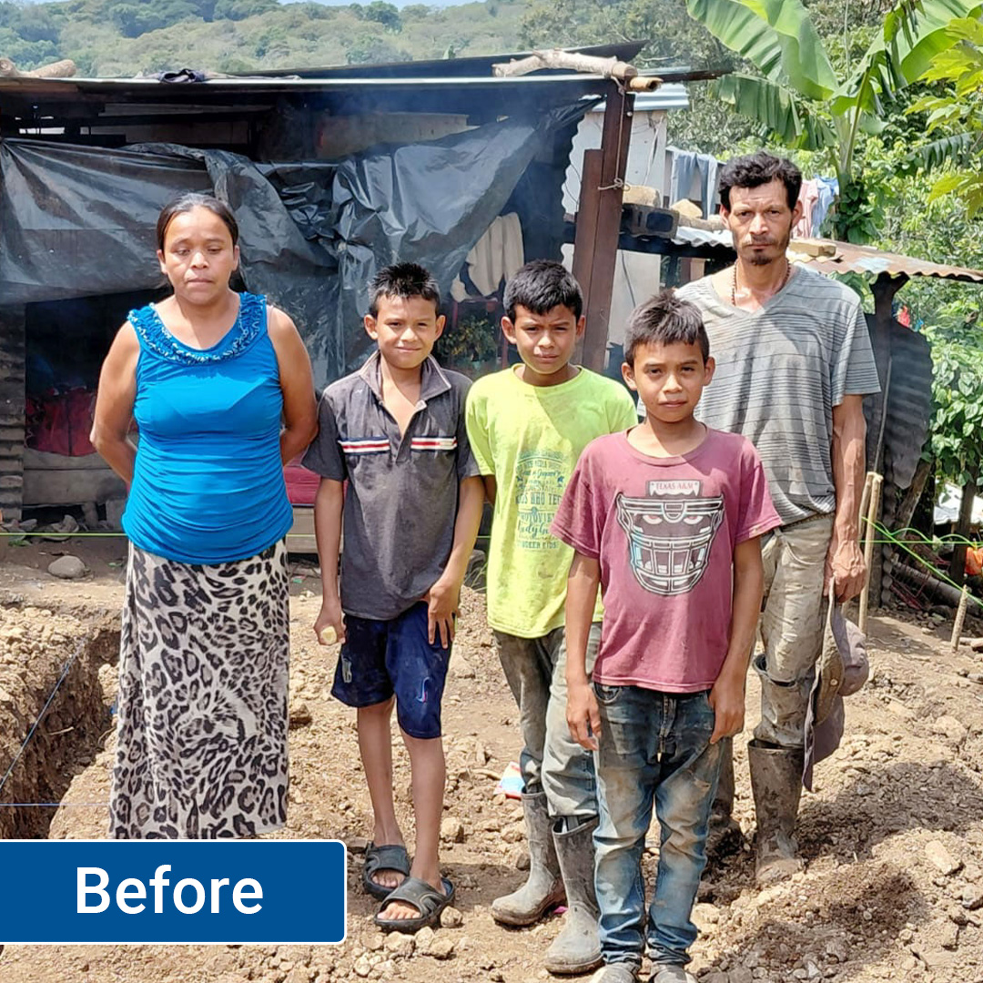 A family stands in front of their dirt-floored, tarp-covered dwelling.