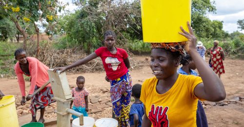 A woman carries a yellow bucket on her head while another woman pumps water from a well. Other women and children stand in the background.