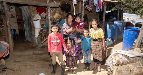 A woman and five young children stand in front of their home made of mud and rusty tin.