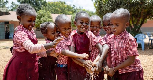 A group of children stands together, holding a water hose.