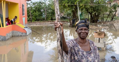 A woman stands in her flooded yard holding onto a tall wooden post.
