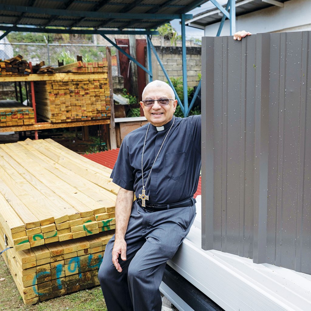 A Catholic bishop holds up a galvanized roof panel while sitting near stacks of lumber and other construction materials.