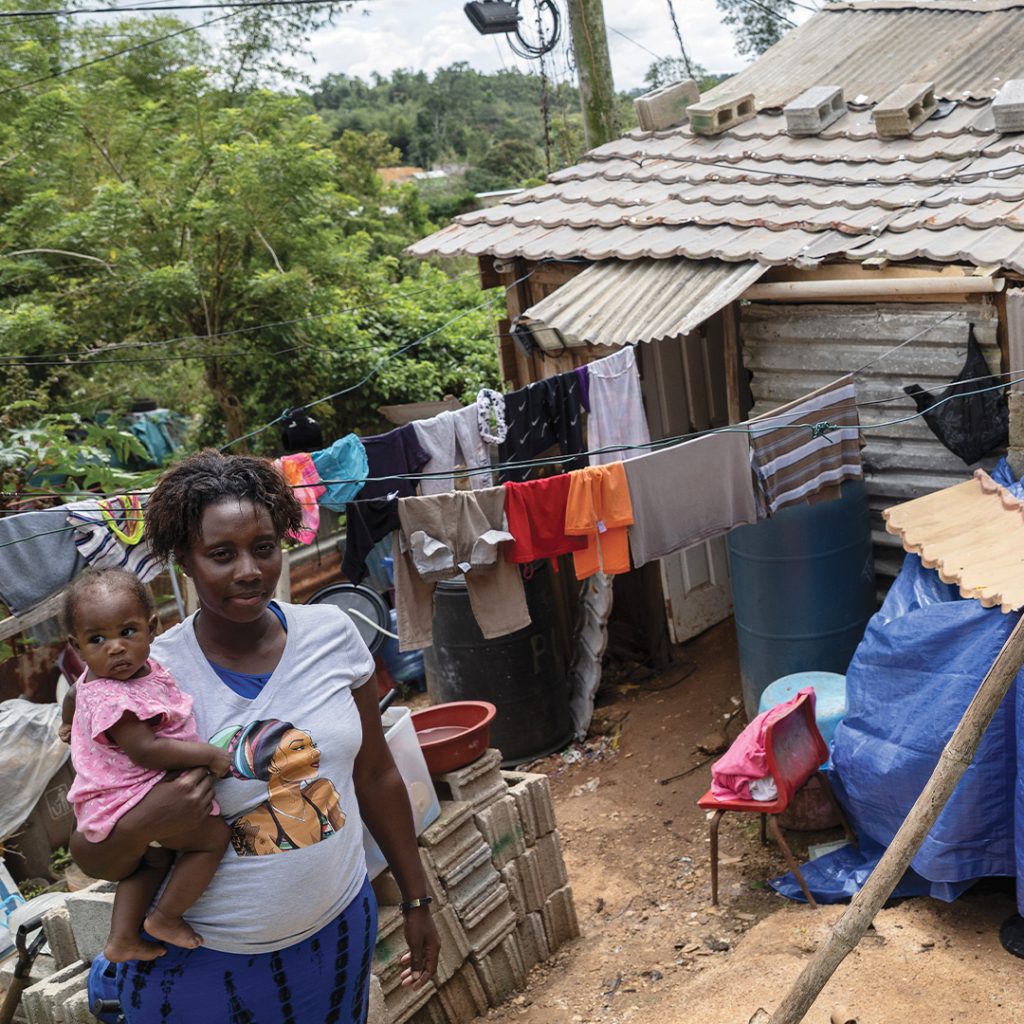 A mother and child stand in the small yard of their makeshift home built with tarps, cinder blocks and other found materials.