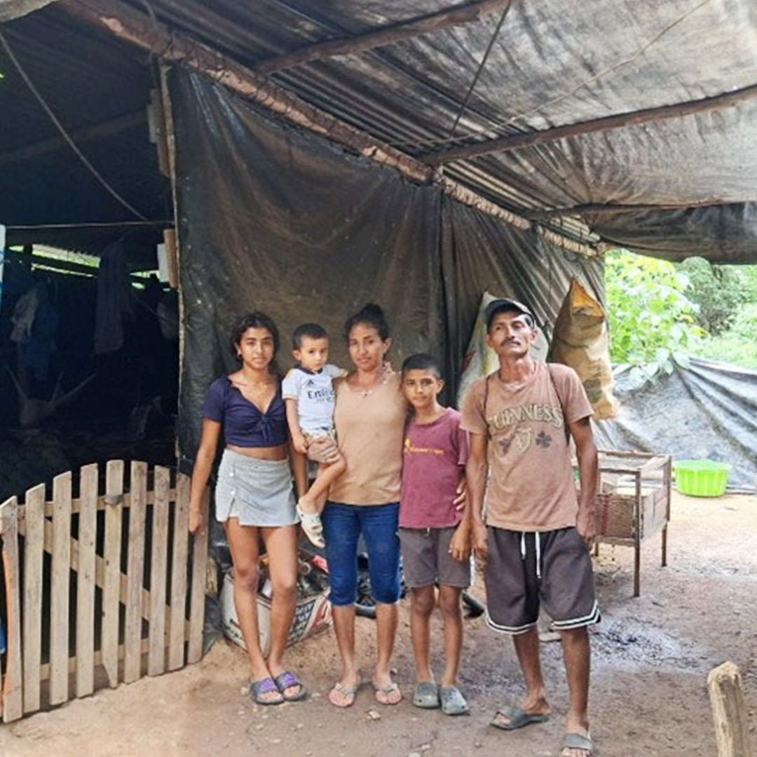 Una familia posa frente a su casa, construida principalmente con lona y tablones de madera.