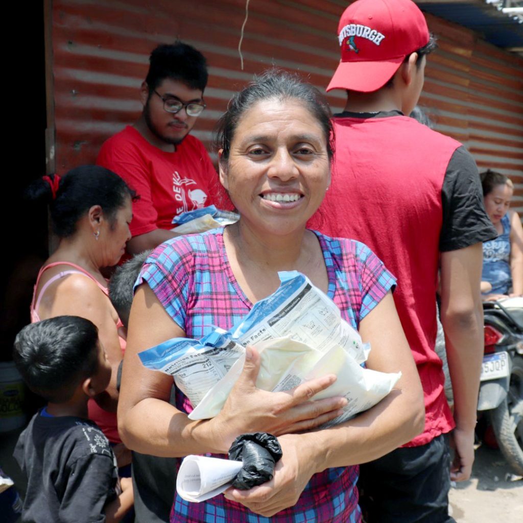 Una mujer sonríe en un evento de distribución de alimentos mientras sostiene bolsas de Vitafood y leche en polvo.