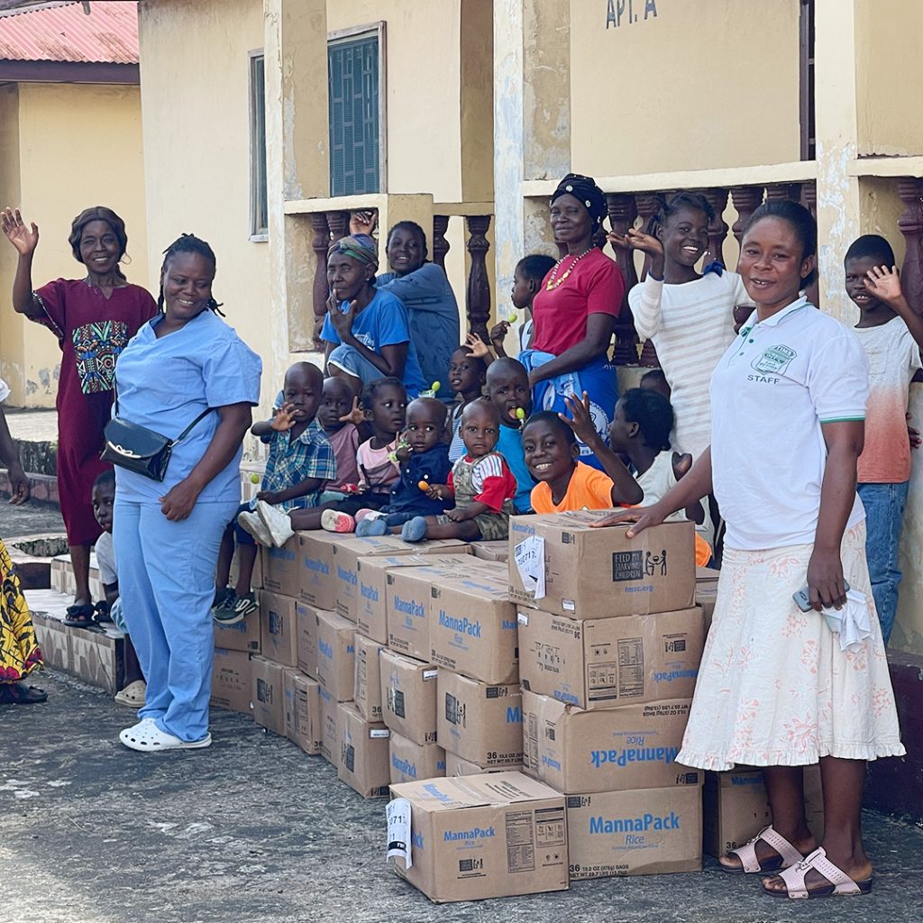 Un grupo de niños y cuidadores sonríen y saludan junto a cajas apiladas de paquetes de comida Vitafood en Liberia.