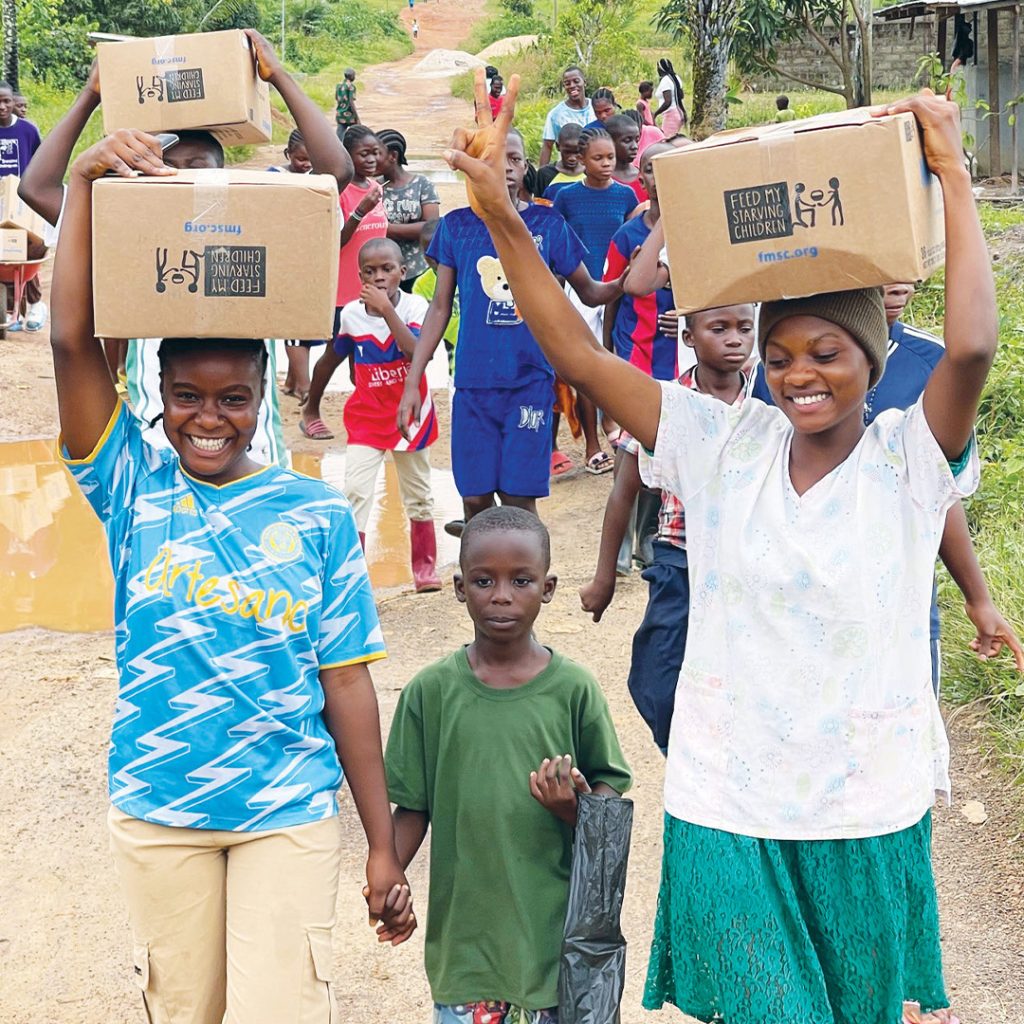 Un grupo de niños y jóvenes carga cajas de cartón de Vitafood por un camino de tierra en Liberia, sonriendo.